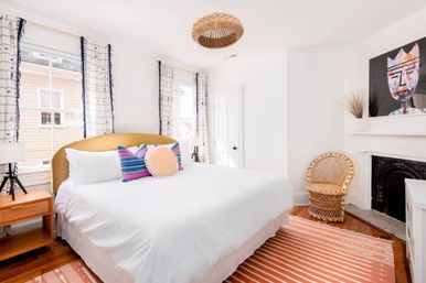 Sunlit modern-boho bedroom with king bed in crisp white linens, mustard upholstered headboard, colorful striped and round throw pillows, rattan wicker chair, coral striped rug, woven pendant light and tall windows.