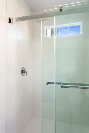 Bright modern bathroom shower with white subway tile, frameless sliding glass door, chrome handle and shower control, and a high horizontal window showing blue sky.