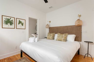 Bright coastal-style bedroom with white linens, woven rattan headboard, patterned throw pillows, hanging wicker bedside lamps, tropical leaf wall art, folded towels on the bed and an open doorway to a bathroom