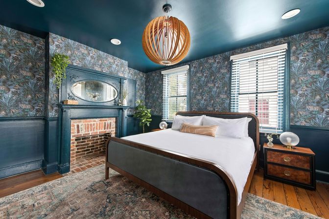 Cozy teal bedroom with floral wallpaper, exposed brick fireplace and oval mirror, large wood-framed bed with crisp white linens, wooden orb pendant, hardwood floors and sunlit windows with blinds