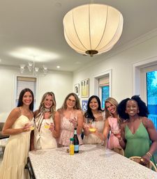 Six women in summer dresses toasting mimosas around a bright modern home kitchen island under a large round pendant light — casual brunch with friends