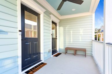 Coastal-style front porch with pastel blue horizontal siding, two black entry doors with welcome mats, a wooden bench, ceiling fan and white railing
