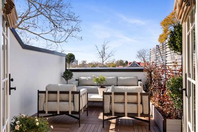 Cozy sunny rooftop terrace with modern outdoor sofa and two armchairs on a wooden deck, potted plants and privacy fence overlooking tree-lined residential rooftops under a clear blue sky.