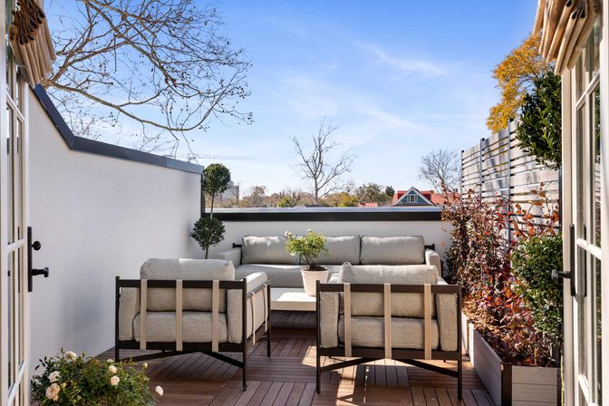 Cozy sunny rooftop terrace with modern outdoor sofa and two armchairs on a wooden deck, potted plants and privacy fence overlooking tree-lined residential rooftops under a clear blue sky.