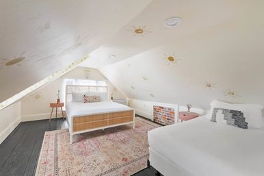 Cozy sun-themed attic loft bedroom with sloped ceilings and sun decals, rattan-front queen bed and white twin bed, pink bedside tables, patterned boho rug, dark hardwood floors and a gable window