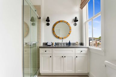 Sunny modern bathroom with white shaker vanity and gray countertop, round wood-accent mirror between black sconces, glass-enclosed shower on the left and a large window with blue sky on the right.