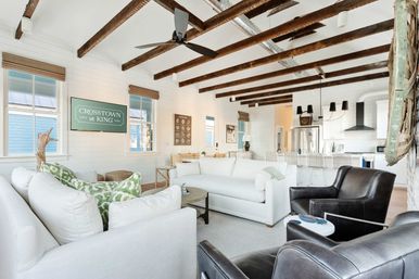 Bright coastal-inspired open-plan living room and kitchen with white sofas, dark leather chairs, exposed wooden beams and bar seating