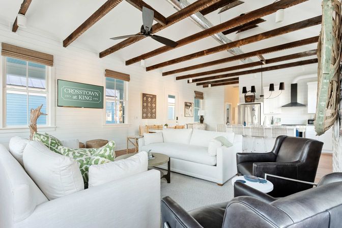 Bright coastal-inspired open-plan living room and kitchen with white sofas, dark leather chairs, exposed wooden beams and bar seating