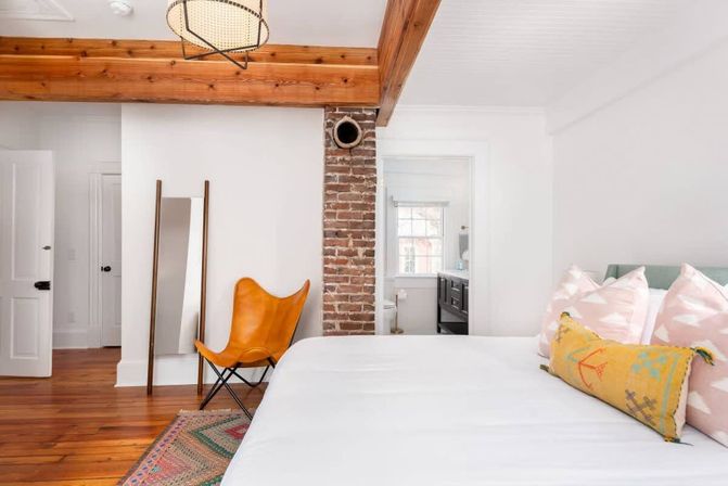 Bright modern bedroom with white bedding, exposed brick column and wooden beam, leather butterfly chair, full-length mirror, colorful rug, hardwood floors and en-suite bathroom.