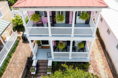 Charming two-story pastel pink house with stacked white porches, hanging fern baskets, black rocking chairs, and a brick driveway