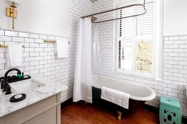 Bright bathroom with white subway tile walls, freestanding black clawfoot tub with white shower curtain and towel, marble vanity with black faucet, brass fixtures, plantation-shutter window, hardwood floor and teal accent stool.