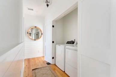Bright coastal farmhouse hallway with hardwood floors and white shiplap walls, a round wood-framed mirror, and a tucked laundry nook with white top-load washer and dryer behind sliding doors.