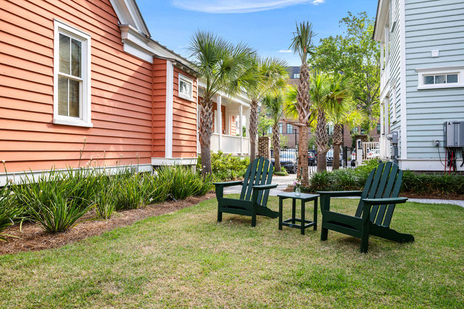 Two dark green Adirondack chairs and a small table on a grassy courtyard framed by palm trees and colorful clapboard houses (salmon and pale blue) in a sunny coastal neighborhood.