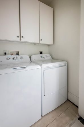 Clean home laundry nook with matching white top-load washer and front-load dryer beneath white wall cabinets on a tiled floor.