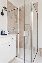 Spa-like modern corner glass shower enclosure with chrome fixtures and beige subway tile, adjacent white vanity with black knobs and blue soap bottle, woven basket on the floor