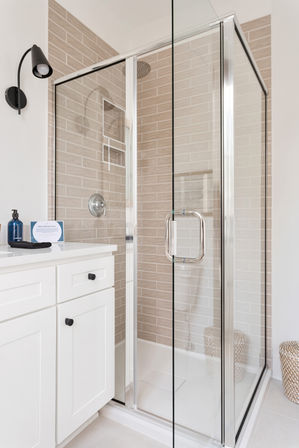 Spa-like modern corner glass shower enclosure with chrome fixtures and beige subway tile, adjacent white vanity with black knobs and blue soap bottle, woven basket on the floor
