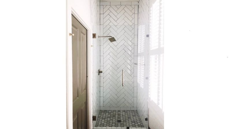 Bright narrow bathroom shower with frameless glass door, white herringbone wall tiles, gray hexagon mosaic floor, square rain showerhead, and shuttered window casting natural light.