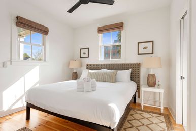 Sunlit urban bedroom with crisp white bedding, woven headboard, hardwood floors, bamboo window shades, bedside lamps and neatly rolled towels.