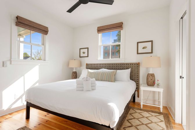 Sunlit urban bedroom with crisp white bedding, woven headboard, hardwood floors, bamboo window shades, bedside lamps and neatly rolled towels.