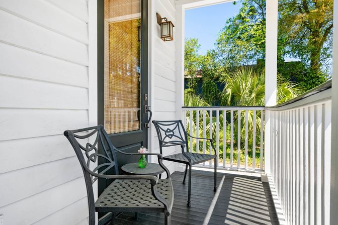Sunlit front porch of a white cottage with two decorative black metal chairs, small round table holding a green vase with flowers, white railing and lush palm trees beyond.