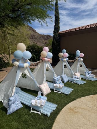Backyard kids’ sleepover setup with white teepee tents topped with pastel blue, pink and cream balloons, inflatable lifebuoy garlands, lounge mats and pillows on green turf, desert mountain and tile-roofed house in the background.