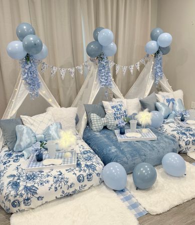 Cozy blue-themed indoor slumber party setup with three mini teepees draped in white fabric, blue balloons, floral garlands, fairy lights and plush pillows.