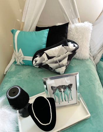Cozy turquoise bedroom nook with white canopy, teal bow-accent pillow, black pillow, geometric diamond-shaped cushion, fluffy white pillow, framed teal high-heel art and a pearl necklace on a black display on a white tray.