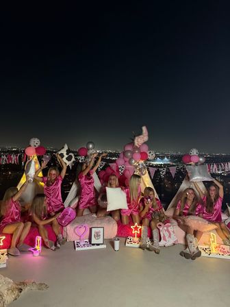 Rooftop nighttime pajama party on a city skyline — women in matching pink satin pajamas laughing and tossing pillows amid pink, silver and cow-print balloons, tents and neon decor.