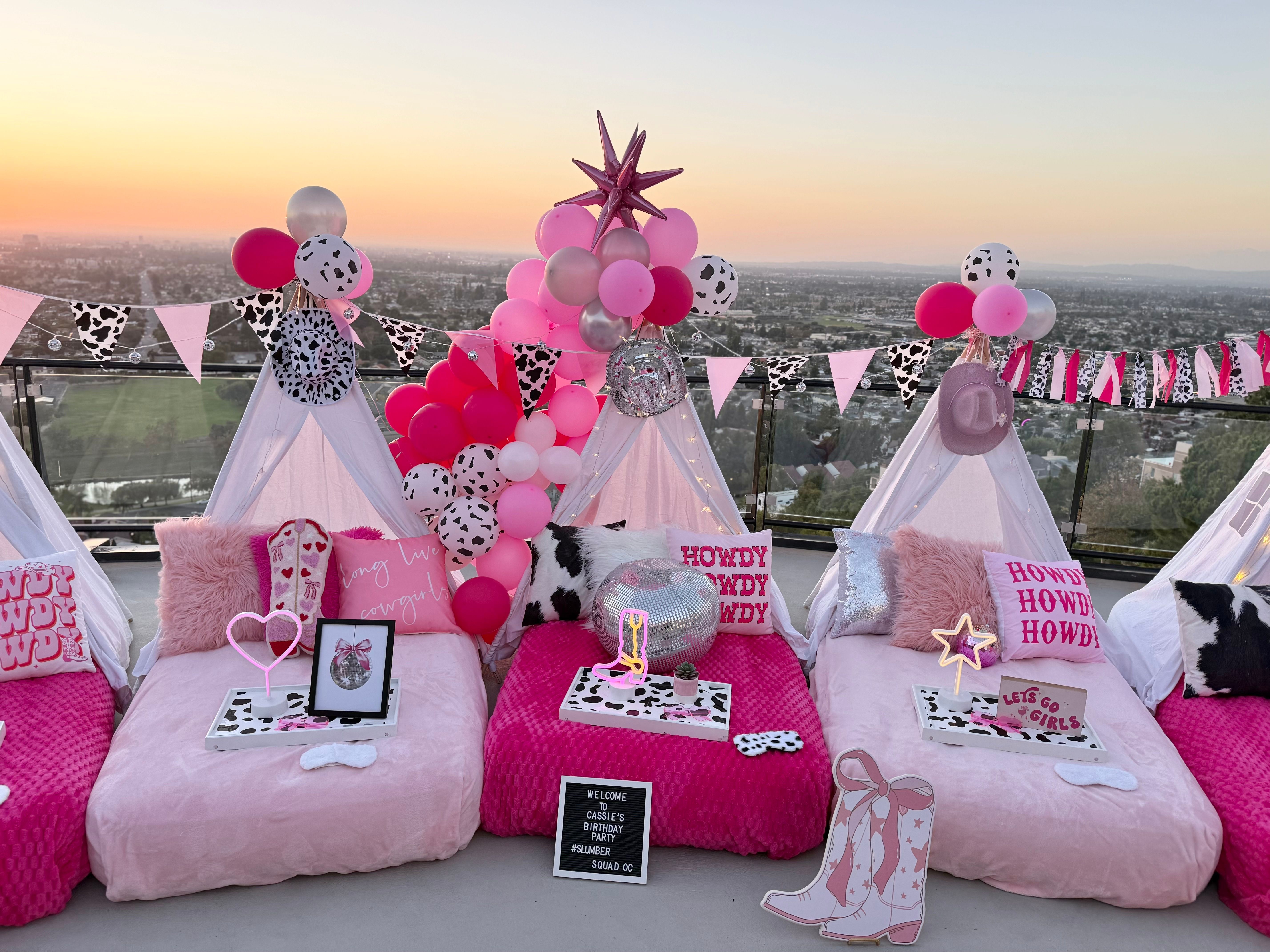 Rooftop sunset slumber party setup with pink mini teepees, cow‑print bunting, pink and silver balloons, disco ball and plush pillows overlooking a city skyline.