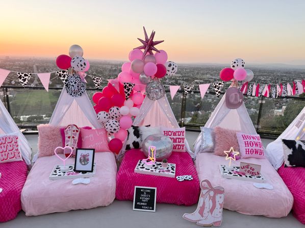 Rooftop sunset slumber party setup with pink mini teepees, cow‑print bunting, pink and silver balloons, disco ball and plush pillows overlooking a city skyline.