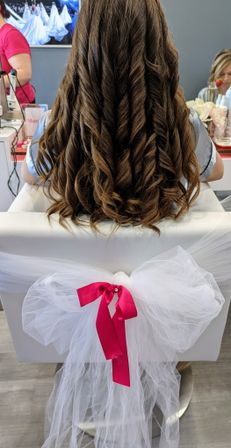 Back view of long brown spiral curls styled for a bridal hairstyle in a salon chair decorated with white tulle and a hot pink bow