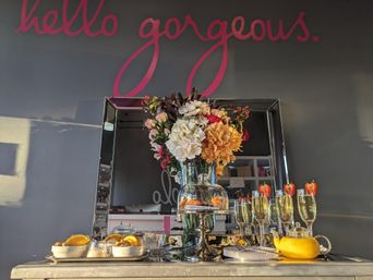 Cheerful reception table with a large mixed-flower bouquet in a vase in front of a mirror, glass cloche of pastries labeled help yourself, champagne flutes topped with strawberries, a yellow teapot and bowls of orange slices under a gray wall painted "hello gorgeous."