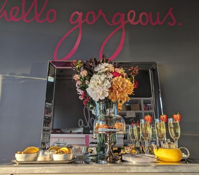Cheerful reception table with a large mixed-flower bouquet in a vase in front of a mirror, glass cloche of pastries labeled help yourself, champagne flutes topped with strawberries, a yellow teapot and bowls of orange slices under a gray wall painted "hello gorgeous."