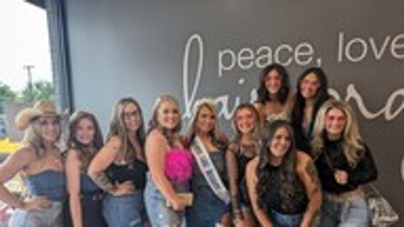 Group photo of a bachelorette party – women in denim and black smiling inside a trendy salon or boutique in front of a gray wall mural reading 'peace, love'.