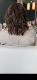 Back view of a shoulder-length wavy brunette lob with textured waves, styled in a modern salon chair with pendant lights and a mirror in the background.