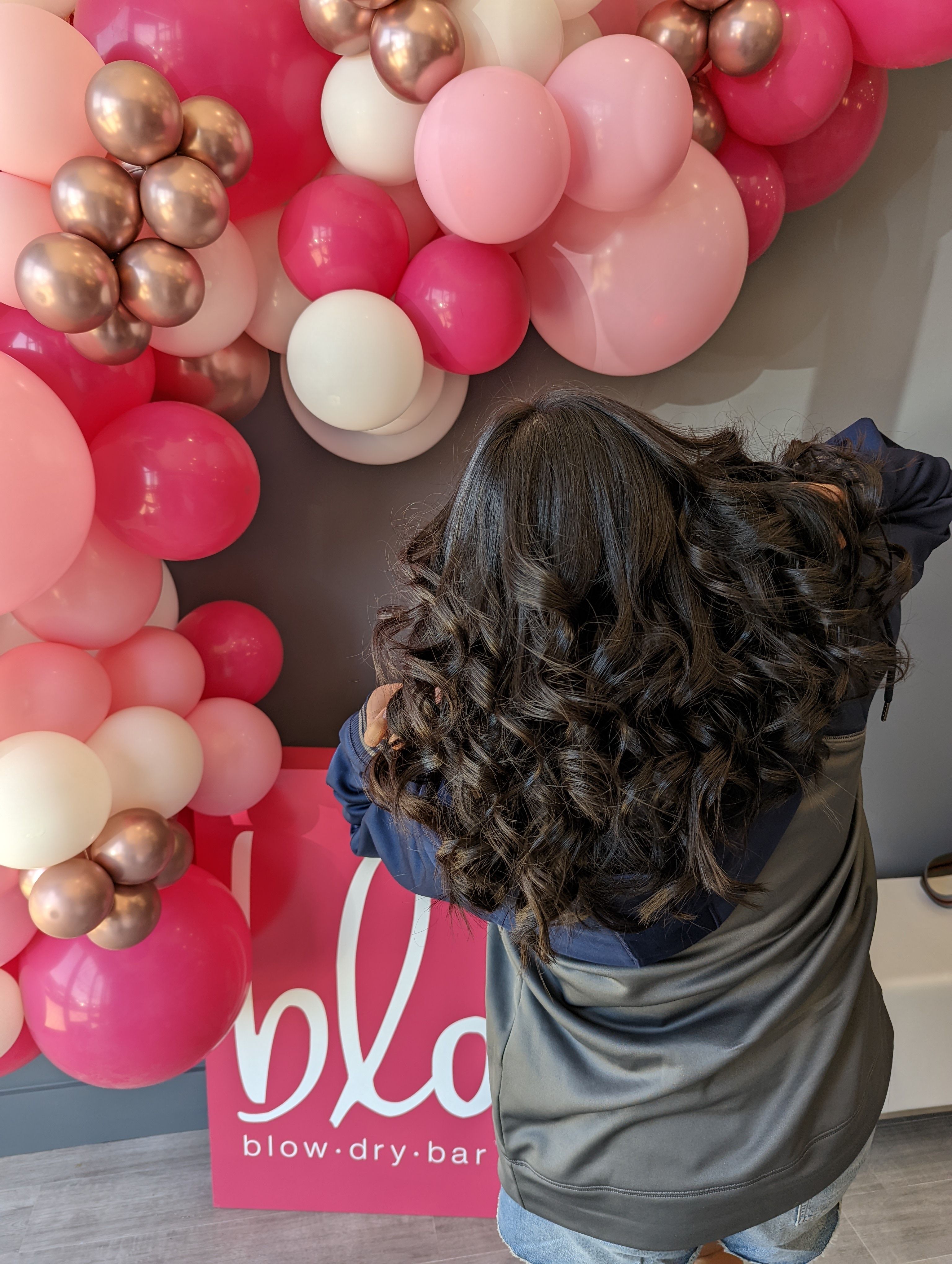 Back view of a person with glossy dark bouncy curls after a salon blowout, posing in front of a pink, white, and rose-gold balloon arch backdrop.
