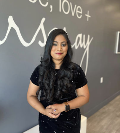 Smiling woman with long glossy black curls wearing a black star-print dress and smartwatch, hands clasped while posing inside a modern salon against a gray wall with white cursive lettering.