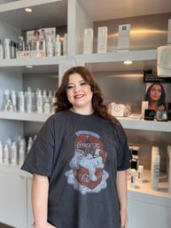 Smiling young woman with styled brown hair and makeup wearing a vintage Coca-Cola tee, posing in a modern hair salon in front of shelves of haircare products