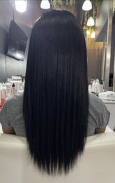 Back view of a client with long, sleek, jet-black hair seated in a salon chair, glossy straightened finish with salon lights, mirror, and styling products visible in the background.