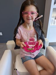 Smiling young girl with wet hair and pink glasses in a kids salon chair holding a glass of water balanced on bright pink rose petals