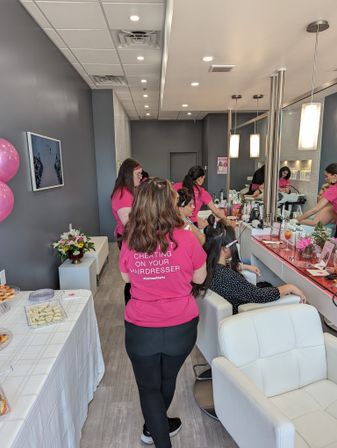 Bright modern hair salon interior with stylists in pink shirts working at mirrored styling stations, white leather chairs, pink balloons and a refreshment table with snacks and flowers
