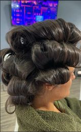 Side profile of a client with dark brown hair in large salon rollers creating glossy, vintage-style barrel curls in a hair salon setting.