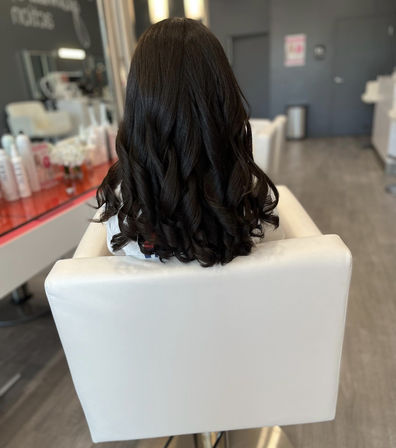Back view of glossy dark brown hair styled in loose spiral curls, seated in a white salon chair inside a modern hair salon with styling products on the counter.