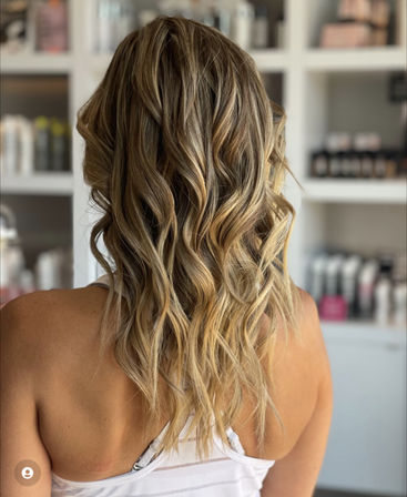 Sun-kissed blonde balayage with loose beachy waves on shoulder-length hair, photographed from the back in a bright salon setting.