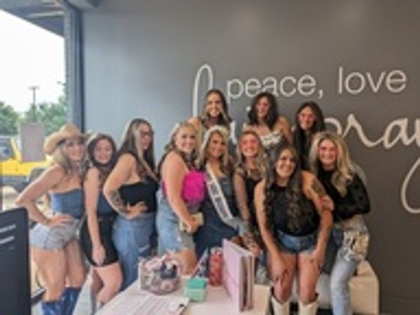 Group of women posing indoors for a bachelorette party, bride-to-be wearing a sash surrounded by friends in denim and boots, pink gifts on a table and a gray wall with 'peace, love' script behind them.