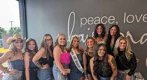 Smiling group of friends pose indoors by a decorative wall sign; wearing denim and black outfits, one woman wears a sash and holds a bright pink bouquet for a celebratory outing.
