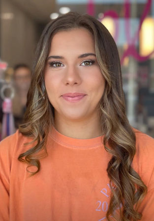 Casual headshot of a smiling young woman with long wavy highlighted brown hair, subtle makeup, wearing an orange t-shirt in a softly lit indoor setting