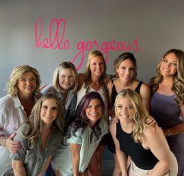 Eight smiling women posing together in a bright beauty studio, group portrait in front of a pink script wall mural reading "hello gorgeous".