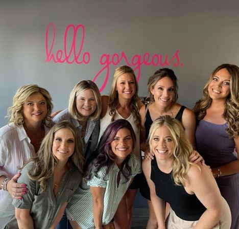 Eight smiling women posing together in a bright beauty studio, group portrait in front of a pink script wall mural reading "hello gorgeous".