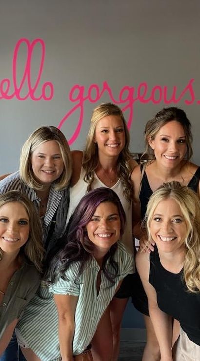 Eight smiling women posing together in a bright beauty studio, group portrait in front of a pink script wall mural reading "hello gorgeous".